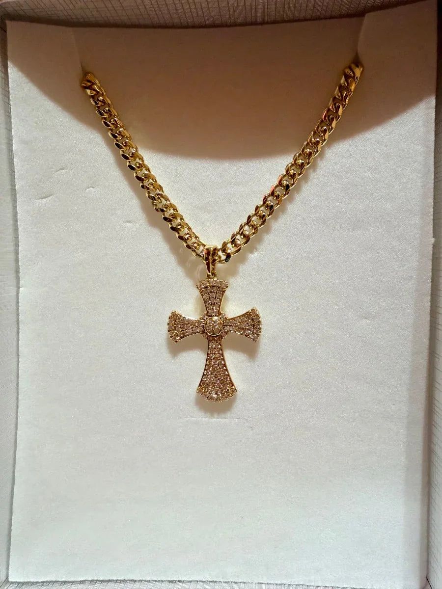 Collar de cadena gruesa dorada con dije de cruz incrustado de brillantes, sobre fondo blanco.