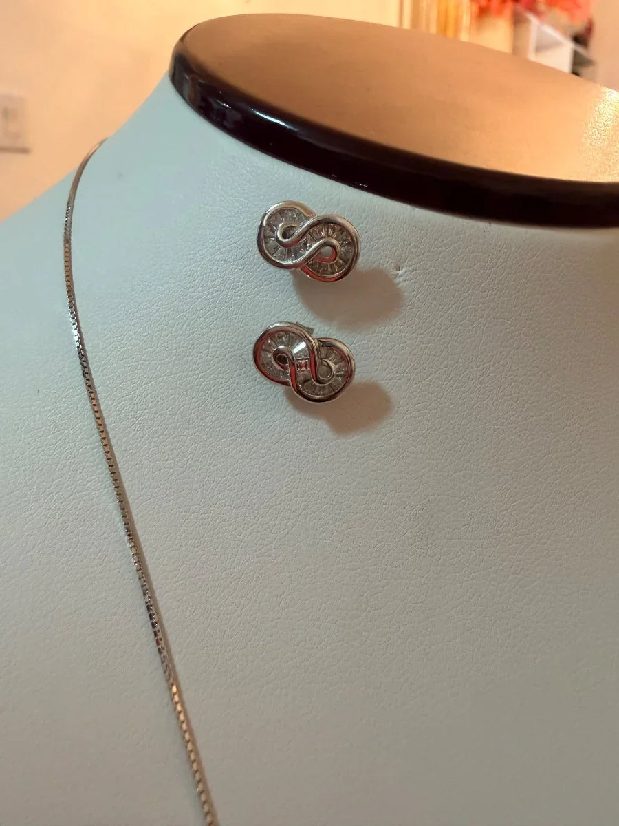 Aretes de plata con diseño de infinito y cadena fina sobre un exhibidor blanco