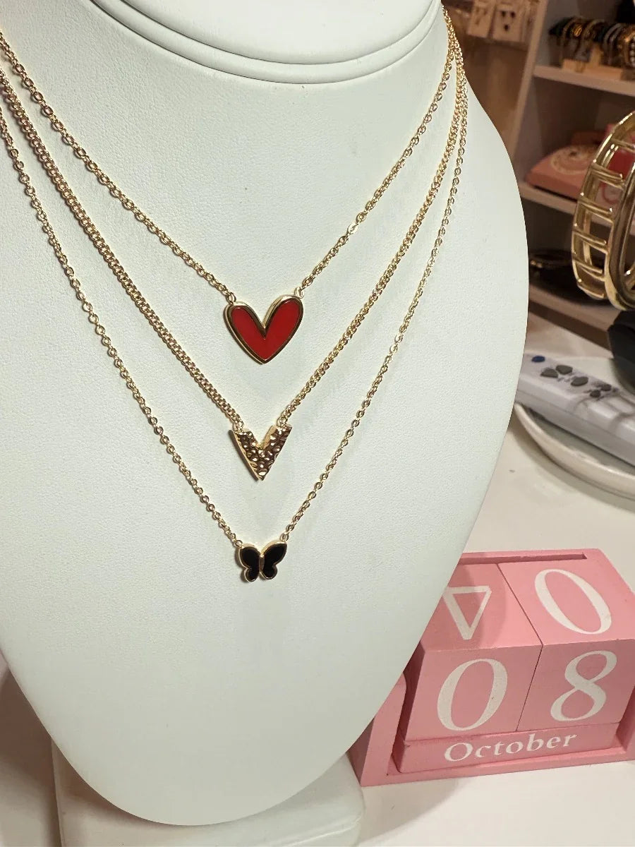 Collares dorados con colgante de corazón rojo, letra V y mariposa negra sobre busto.