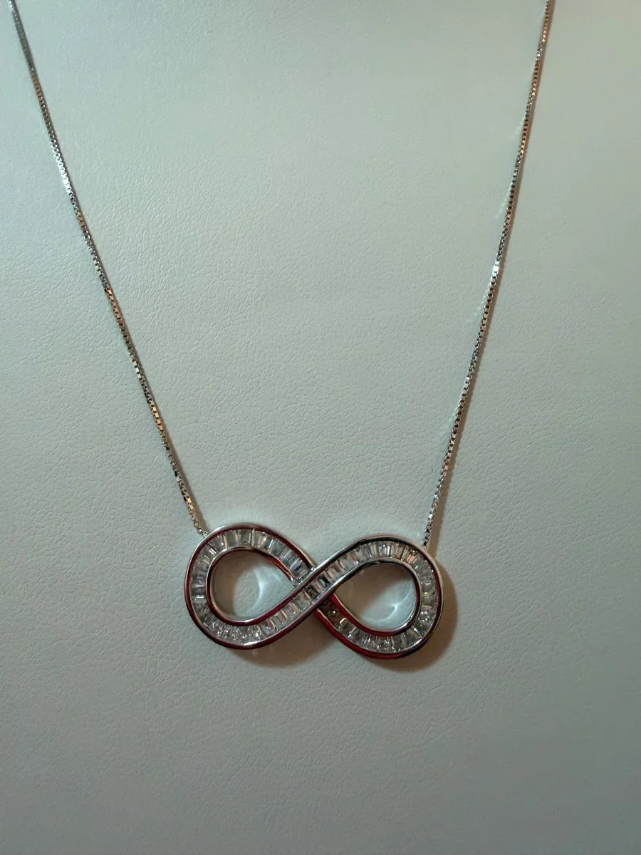 Collar de plata con dije de infinito decorado con piedras brillantes, fondo neutro.