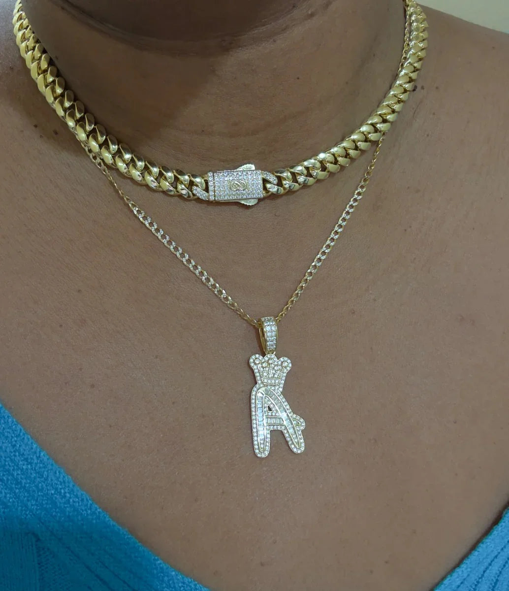 Collar grueso dorado y cadena fina con dije de letra A coronada con circonias, sobre piel morena
