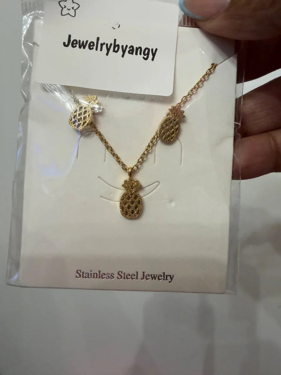 Conjunto de collar y aretes de piña dorados en acero inoxidable sobre fondo blanco.